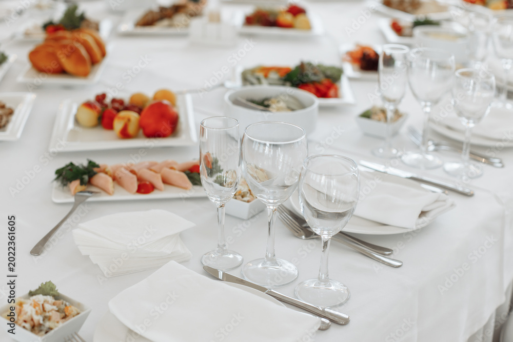 Fototapeta premium Close-up of a formal place setting with clear wine glasses, a white folded napkin, and plates of fresh appetizers on a banquet table.