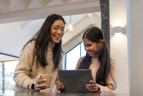 African American mom leaning with watch holding mug, Indian daughter child using tablet at kitchen