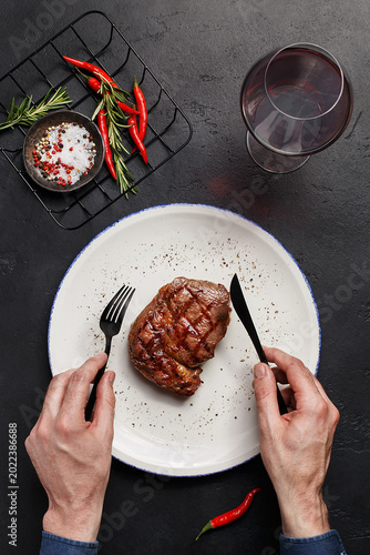 Men's hands holding fork and knife over plate with grilled ribeye steak on black stone background with glass of red wine, chili peppers, salt and rosemary. Overhead view, flat lay