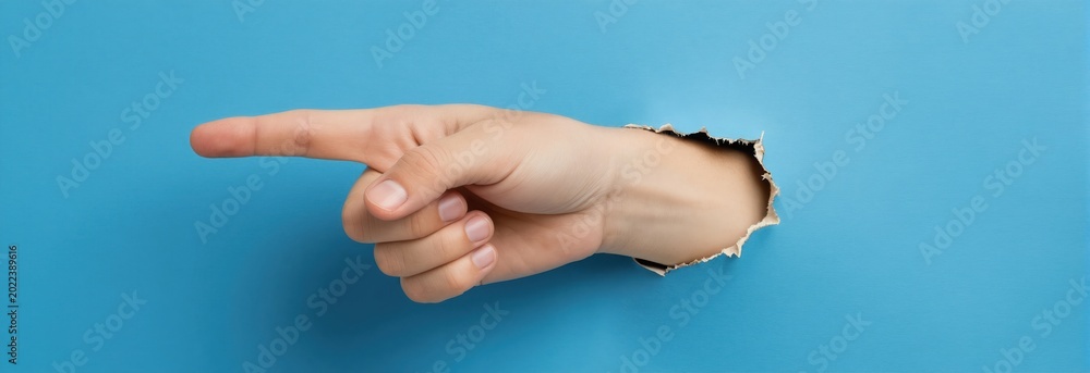 Fototapeta premium Hand points to the right through a torn hole in a blue paper background. Index finger extended shows direction or indicates something important. Copy space available.