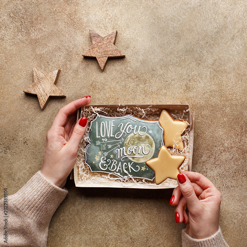 Foto Female hands in sweater holding cardboard gift box with decorated gingerbread cookies in shape of stars and with Love you to the moon and back text over beige concrete background