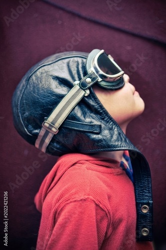 Side profile of a boy in a red sweater and leather aviator helmet with goggles, against a textured dark red background. Nostalgic and playful vibes.