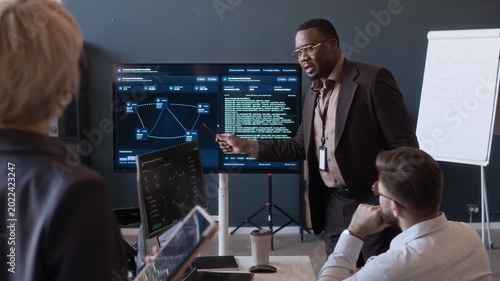 Medium long shot of young Black tech specialist presenting network data on screen to cybersecurity team while meeting in office