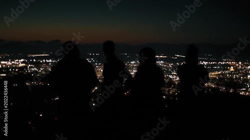 Group of tourists on viewing platform on mountain admire and take pictures on their mobile phones of panorama of night city with many lights
