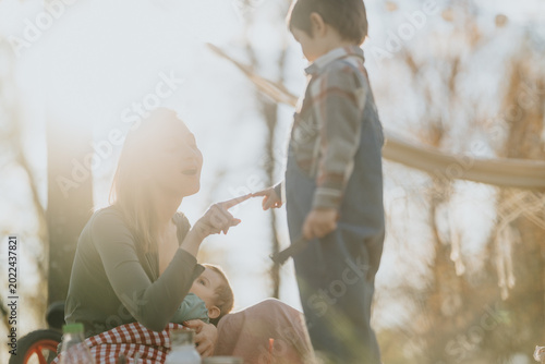A mother sits outdoors holding a baby while interacting with her older child. Warm backlit scene shows family bonding during a casual outdoor moment.
