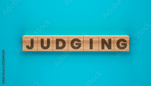 Slika na platnu Judging Word Blocks: A close-up shot of the word JUDGING spelled out with wooden blocks set against a vibrant turquoise backdrop