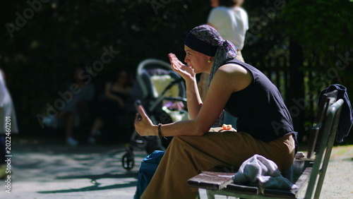 Woman undergoing chemotherapy on park bench during video call, smiling while listening and reacting with expressive gesture in warm sunlight