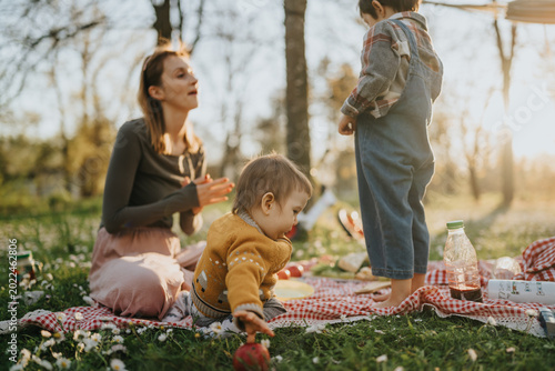 A mother sits with two young children having a picnic on a checkered blanket in a park. Soft sunlight filters through trees as the family relaxes and the children play on the grass.
