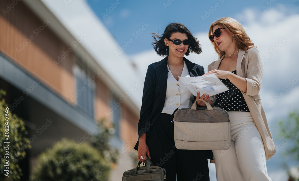 Fototapeta premium Two businesswomen review documents together outdoors. Colleagues consult papers while walking near a modern office building.