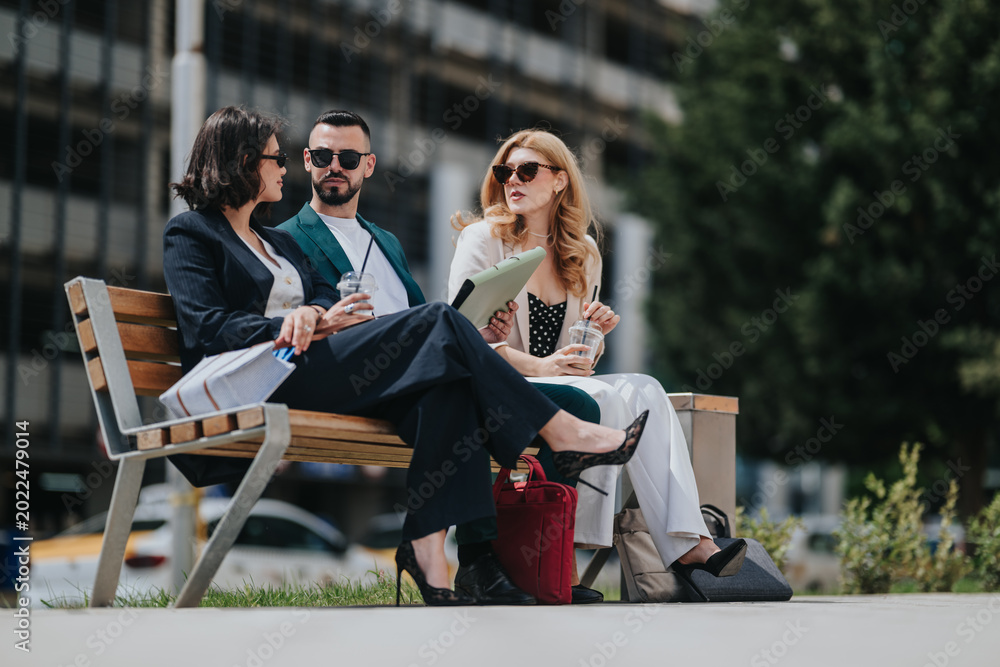 Fototapeta premium Three businesspeople sit on a bench discussing documents and using tablets and drinks. An informal outdoor meeting unfolds in an urban setting as colleagues review papers together.