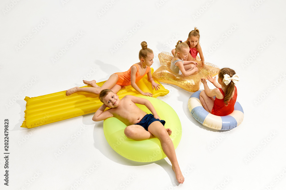 Obraz premium Children relaxing on colorful inflatable pool floats in white studio with large copy space. Concept of summer camp promo, kids swimwear ads, family travel offers and pool toy marketing.