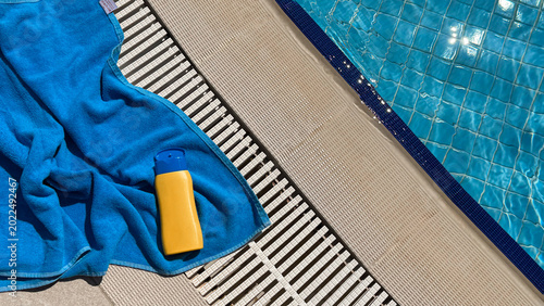 Blue towel and yellow sunscreen bottle placed beside a swimming pool with clear water and tiled surface reflecting sunlight on a warm day