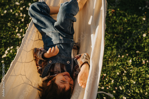 A young child lies in a hammock wearing denim overalls and a plaid shirt, smiling with eyes closed. Sunlit grass and scattered small flowers surround the hammock as the child relaxes playfully.