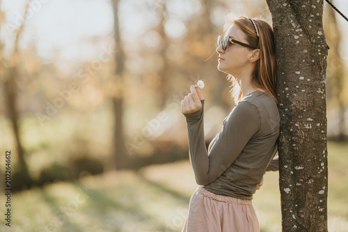 Tableau sur toile Young woman wearing sunglasses leans against a tree holding a small daisy