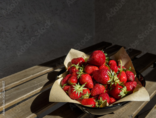 Bowl of fresh strawberries on wooden table in sunlight. Concept of summer harvest, healthy eating, rustic food styling and seasonal freshness