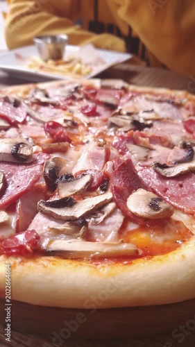 Close-up view of a freshly baked pizza topped with mushrooms, ham, and salami pieces on a wooden board. Concept of Italian cuisine, junk food addiction, and savory fast food indulgence.