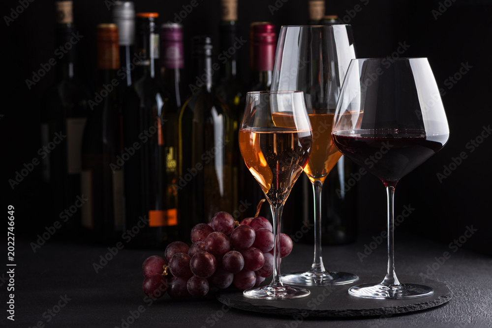 Fototapeta premium Three glasses of red, white and sparkling rose wine with bunch of grapes on dark slate background