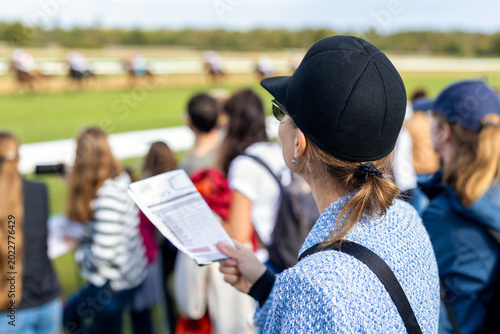 Billede på lærred Elegant woman watching horse racing event while holding betting program near racetrack crowd