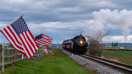 A steam train moves along railroad tracks in a countryside setting. American flags are planted along the route, and there are clouds overhead. The scene is lively with movement and nature.