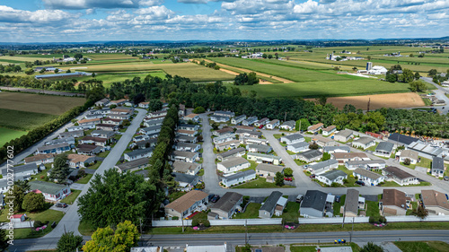 A suburban area with rows of mobile, manufactured homes. Green fields stretch into the background under a partly cloudy sky. The image captures a simple residential neighborhood.