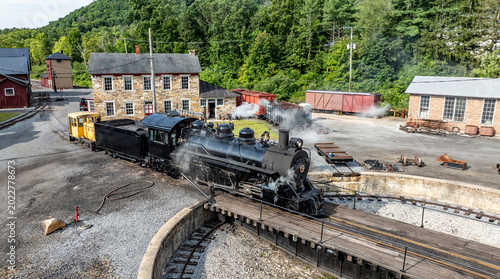 Photos A steam locomotive moves on a circular turntable at a train yard surrounded by green trees and buildings
