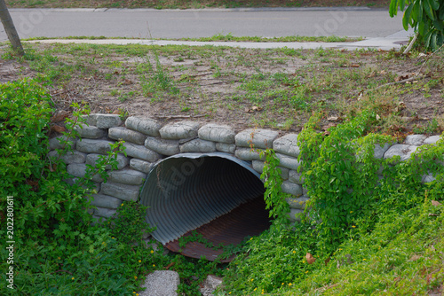 An overgrown corrugated metal culvert and drainage pipe set into a sandbag retaining wall, surrounded by dense grass, vines and small yellow flowers conveying nature reclaiming infrastructure.
