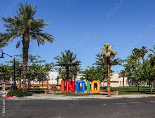 Indio, California, USA - April 15, 2026: Colorful name sign along road on side of green park with plenty of palm and other trees under blue sky