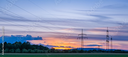 High-voltage transmission towers silhouetted against sunset sky, power lines crossing rural field, Sustainable energy infrastructure, green crops, distant hills, industrial infrastructure meets nature