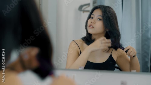 Samiling happy Asian woman wearing a black dress brushing her hair with hairbrush, looking at mirror sitting in front of the dressing table at home. Female haircare and cosmetics concept.
