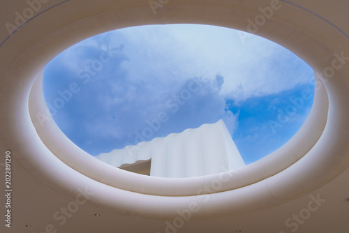 Modern architecture with circular skylight and white wavy wall under blue sky with clouds, bright and airy design