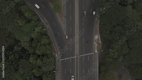 Top down aerial view of vehicles driving on a multi lane highway surrounded by green trees in the city