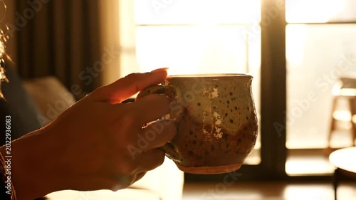 Person holding ceramic mug by sunny window in morning light