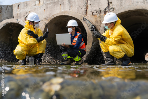 Team of environmental scientists in hazmat suits and safety gear collecting water samples and recording data at a drainage pipe for pollution analysis.