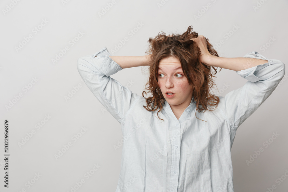 Naklejka premium Surprised young woman with curly hair in white shirt holds head with hands, looking to side. Concept of shock, confusion, and unexpected news in casual studio setting.