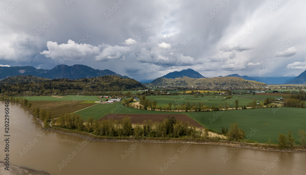 Fototapeta premium Aerial View Of Fraser Valley River, Farmland And Mountains Under Dramatic Cloudy Sky In British Columbia