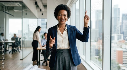 A smiling Black woman stands confidently in a modern office. She wears a navy blue blazer over a white shirt and gray skirt.