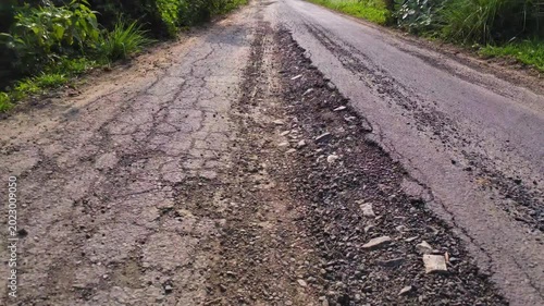 Damaged Asphalt Road with Potholes in Rural India Transportation Infrastructure Close Up. Rural Indian Road with Broken Asphalt Surface and Deep Cracks Slow Motion. Cracks and Road Potholes Outdoors.