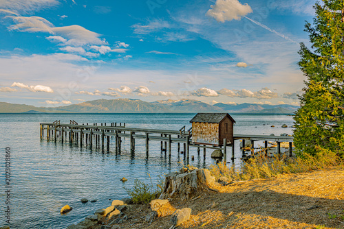 Valokuva A weathered wooden dock with a cedar-shingled boathouse extends over Lake Tahoe's blue water, with snow-dusted Sierra Nevada peaks rising across the horizon