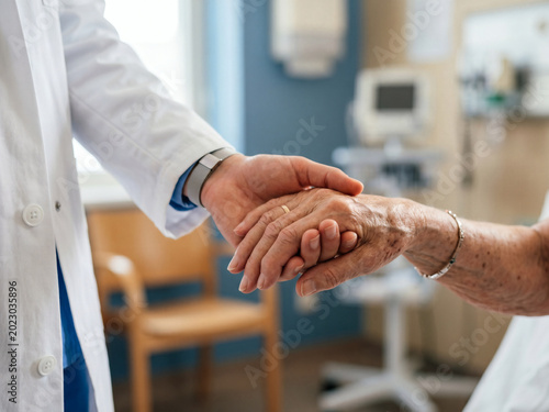 Caregiver holds elderly hand with white coat sleeve in clinic room