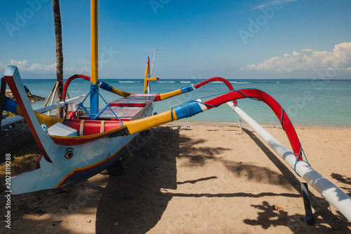 Jukung or kano - traditional fishing boat on Sanur beach in Bali beside calm tropical sea beneath bright blue sky. Scenic island coastline, seaside travel and Indonesian coastal lifestyle