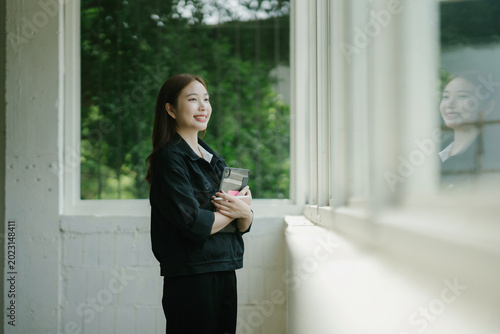 Smiling asian woman holding a tablet by a window, finding inspiration and feeling productive in a modern office