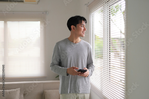 Smiling young Asian man standing by a window with blinds, holding a smartphone and looking outside, enjoying privacy at home