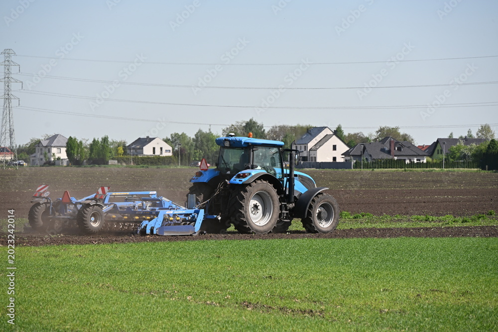 Obraz premium Blue Tractor with Harrow Working on Field Under Sunny Sky with Contrast of Green and Plowed Soil