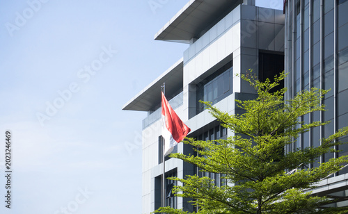 low angle view of the indonesian national flag waving in front of a modern office building with silver metallic cladding and green trees against a clear blue sky