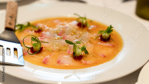 Thinly sliced fish served with citrus dressing, garnished with microgreens and decorative sauce dots. Plate and fork visible on white table.