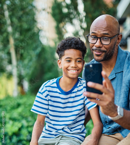 Portrait of father and son having fun using a mobile phone and taking  a selfie photo with camera outdoors in park or nature,, family life, parenting, love and bonding concepts