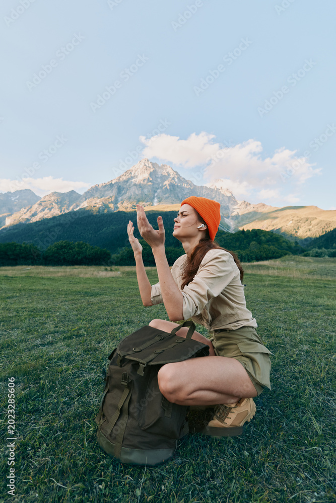 Fototapeta premium Woman kneeling in a grassy field with distant mountains, wearing an orange beanie and backpack. A candid outdoor lifestyle shot celebrating nature, exploration, and adventure.