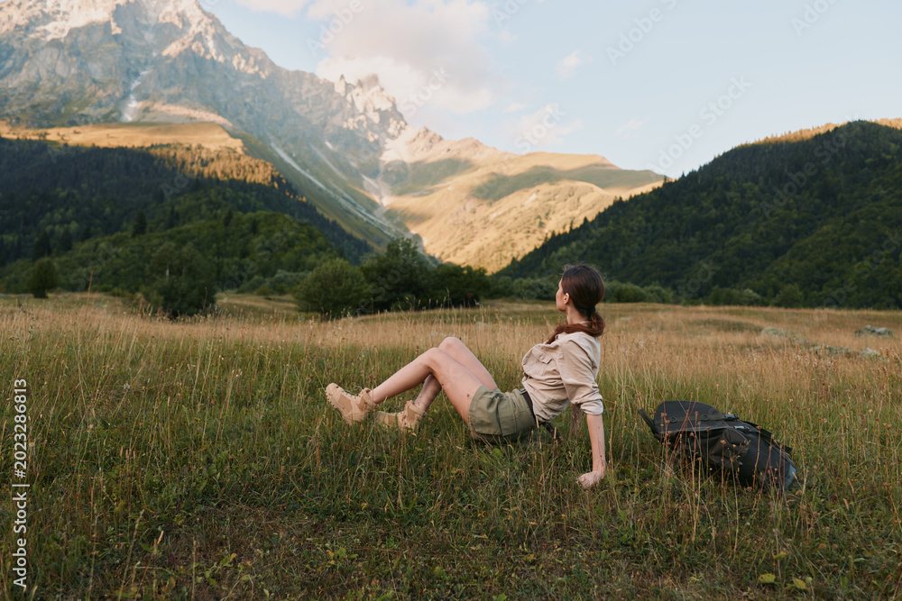 Fototapeta premium A woman rests in a sunlit meadow with distant mountains, backpack beside her. A calm moment of outdoor life, hiking, and nature, inviting exploration and solitude.