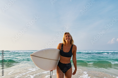 Canvas Print Smiling woman with surfboard stands on sandy beach
