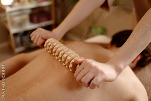 Close up of young adult man receiving wooden roller massage on back, supporting muscle recovery and body care. Suitable for spa wellness, physiotherapy, relaxation marketing
